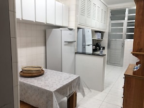 Uma cozinha pequena com armários brancos, uma geladeira, uma cafeteira e uma mesa com toalha de mesa florida.