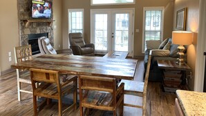 A spacious living room with a large wooden table, comfortable seating, a stone fireplace, and a television mounted on the wall. - Carr's Northside Motel and Cottages (Gatlinburg)