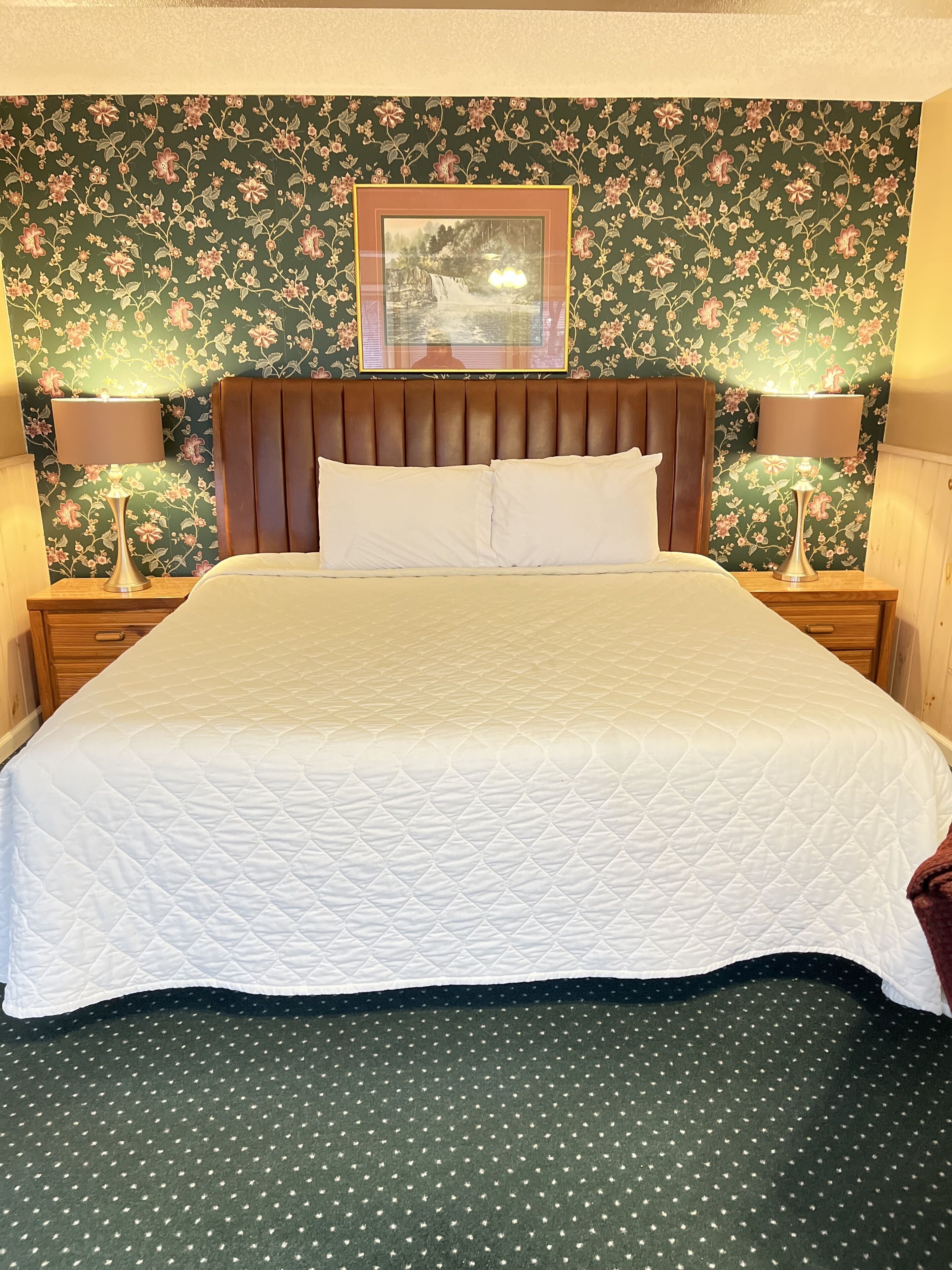 A neatly made bed with a quilted white cover, flanked by two nightstands with lamps, against a floral wallpaper backdrop.