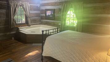 A log cabin room with a wooden floor, a bed with a quilt, a window with curtains, and a framed picture on the wall.