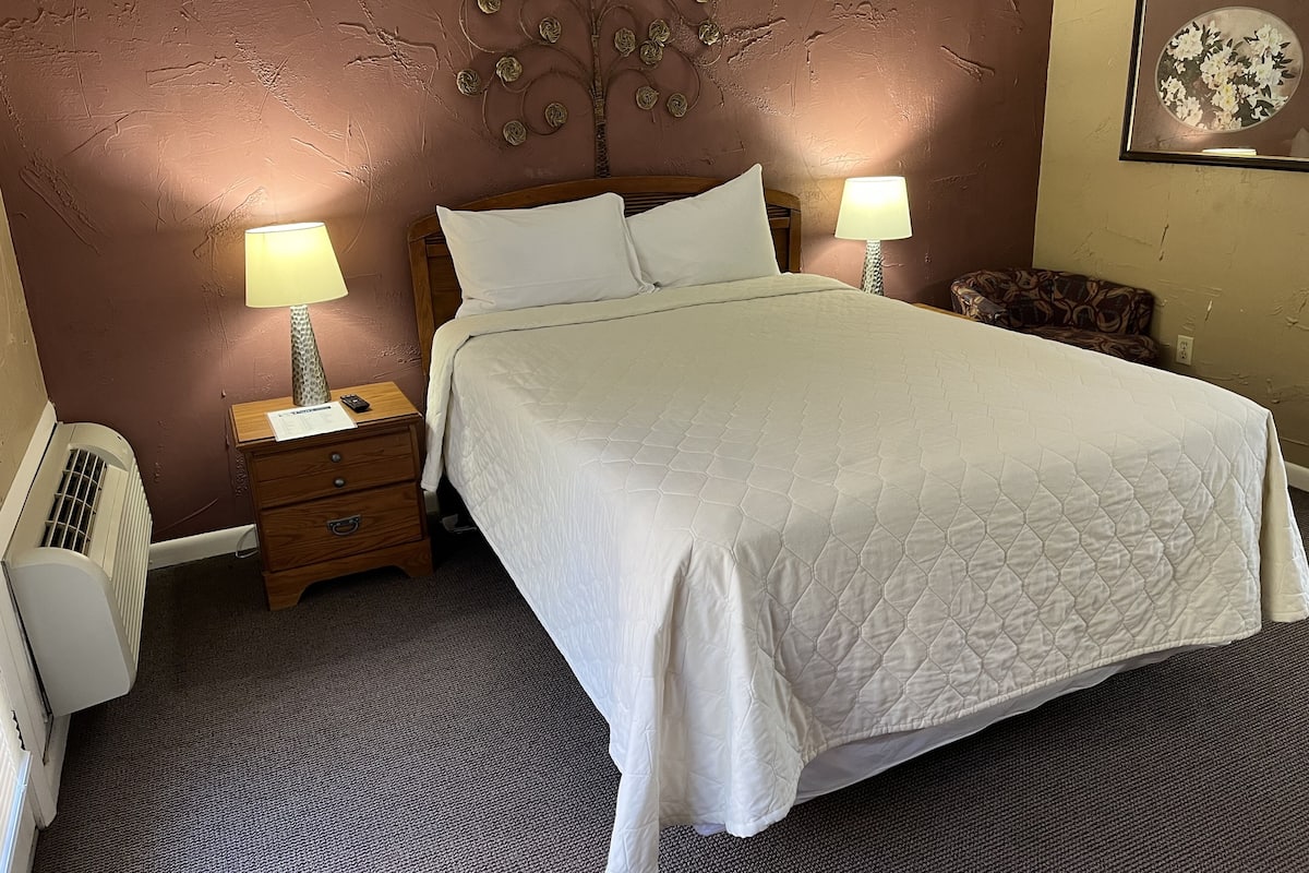 Een net opgemaakt bed met witte beddengoed, twee nachtlampjes, een houten nachtkastje, een radiator aan de muur en een lijst met bloemistisch kunstwerk.