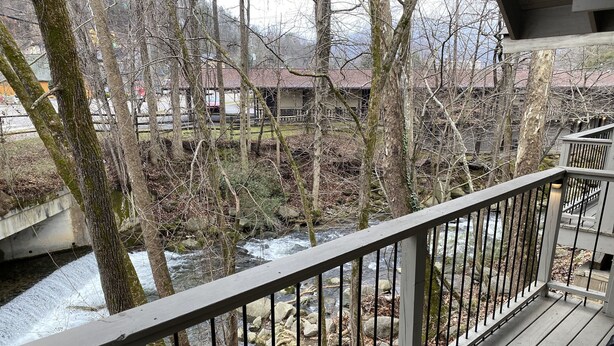 A wooden deck with a metal railing overlooking a river with trees and buildings in the background. - Carr's Northside Motel and Cottages (Gatlinburg)