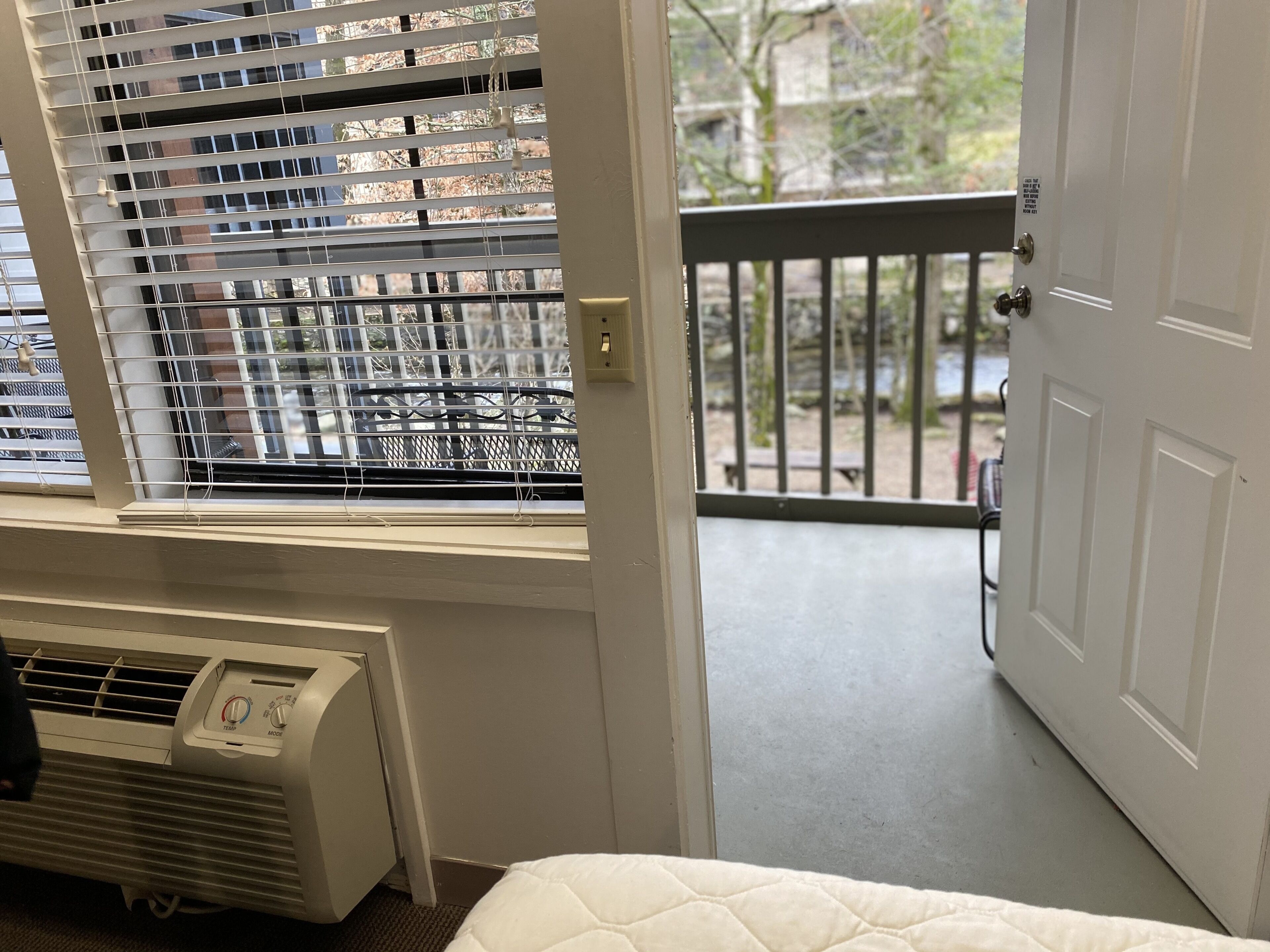 Una habitación con radiador, ventana con persianas, puerta y vista al balcón exterior.