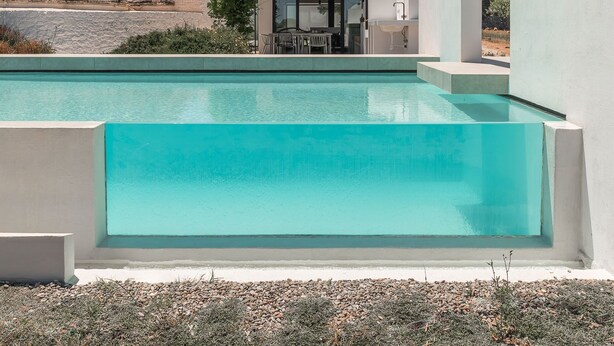 Un área de piscina moderna con una piscina azul cristalina, un edificio blanco con techo plano y una estructura de piedra al fondo.
