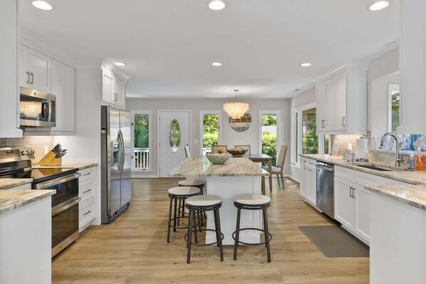 A modern kitchen with white cabinetry, stainless steel appliances, a marble island with stools, and a chandelier.