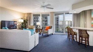 Ein Hotelzimmer mit Balkon, Deckenventilator, Fernseher, Sofa, Stuhl und einem kleinen Tisch mit Lampe.