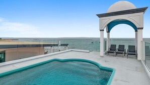 A rooftop pool area with a clear view of the ocean, a covered seating area, and a glass balustrade.