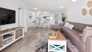 A modern living room with a flat-screen TV, a wooden coffee table, a beige sofa, and a dining area with white chairs and a kitchen in the background.