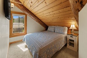 A bedroom with a sloped ceiling, a bed with patterned bedding, a nightstand with a lamp, and a window with blinds.