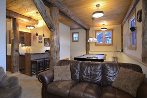 A cozy living room with a pool table, leather sofa, wooden beams, and a kitchen area.