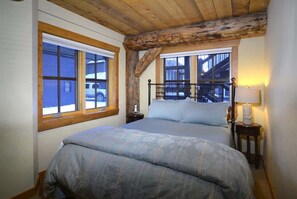 A bedroom with a large bed, wooden beams, a window with a view of a building, and a nightstand with a lamp.