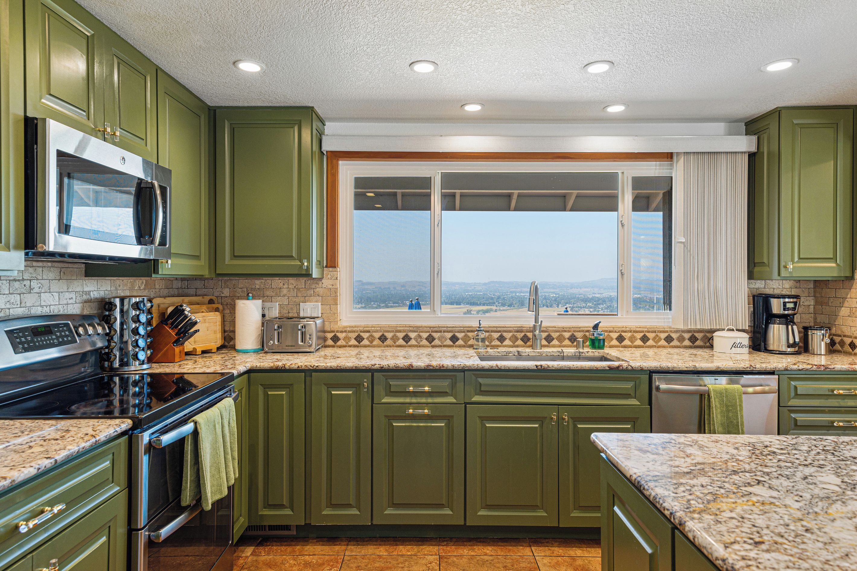 Una cocina con armarios verdes, microondas, estufa, ventana con vistas y cafetera.