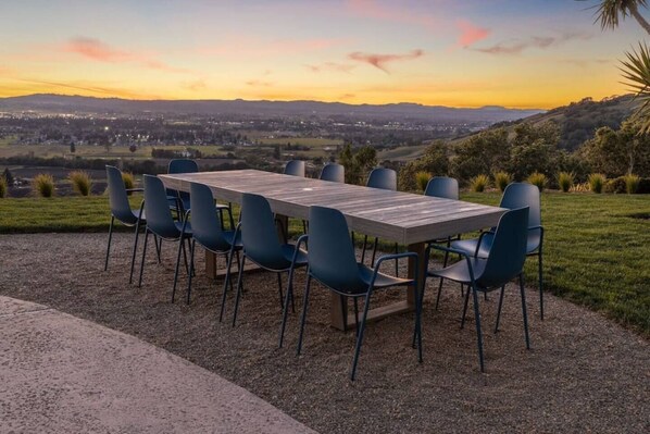 Un área de comedor al aire libre con una mesa larga y sillas, frente a un atardecer sobre un paisaje montañoso.