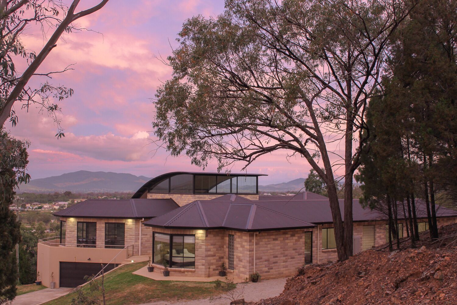 A modern house with a curved glass facade, surrounded by trees and a mountainous backdrop.