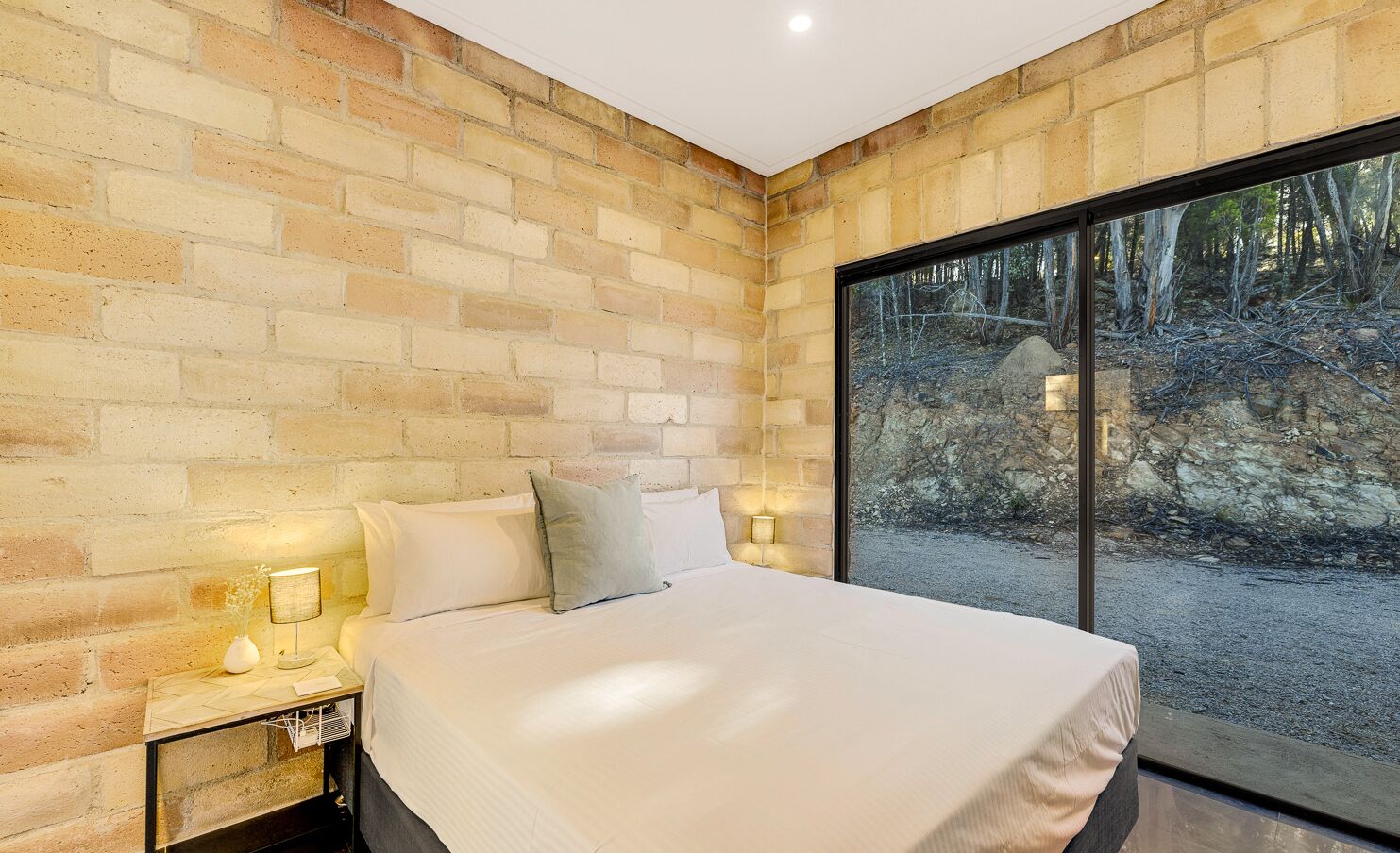 Un dormitorio con pared de ladrillo, una cama grande, lámparas de noche y vistas a un bosque a través de un ventanal.