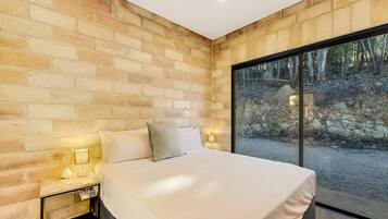 Un dormitorio con pared de ladrillo, una cama grande, lámparas de noche y vistas a un bosque a través de un ventanal.
