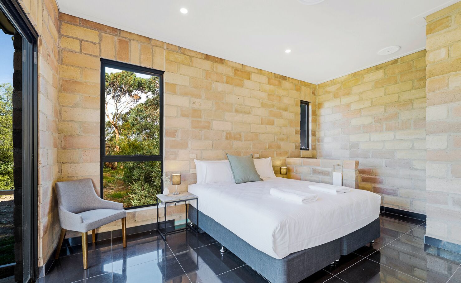 A modern bedroom with a large bed, a chair, a small table, and a window with a view of greenery.