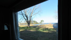 A room with a window overlooking a grassy area with a tree and a body of water. - Little Bay Waterfront Retreat (White Stone)
