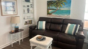 A living room with a dark brown leather sofa, a small white wicker table, and a framed picture of a beach scene.