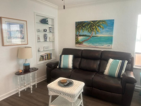Una sala de estar con un sofá de cuero marrón oscuro, una mesita de mimbre blanca y un cuadro enmarcado con una escena de playa.