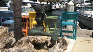 A marina with boats docked, a statue of a mermaid, and colorful chairs.