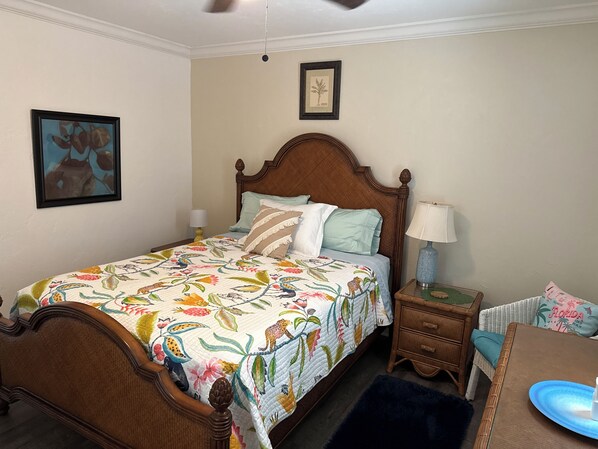 A bedroom with a wooden bed, a nightstand with a lamp, a wicker chair, and a framed plant on the wall.