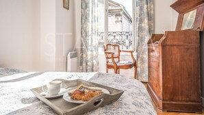 Quarto de hotel com cama, cómoda de madeira, cadeira, varanda com cortinas e bandeja com chávena de café e pastelaria.