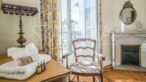 Quarto com cadeira de madeira, mesa com toalhas e amenities, luminária, janela com cortinas, lareira e espelho na parede.