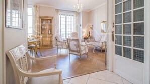 Uma sala de estar espaçosa com um candelabro, uma área de jantar com mesa e cadeiras, e uma janela grande com cortinas.