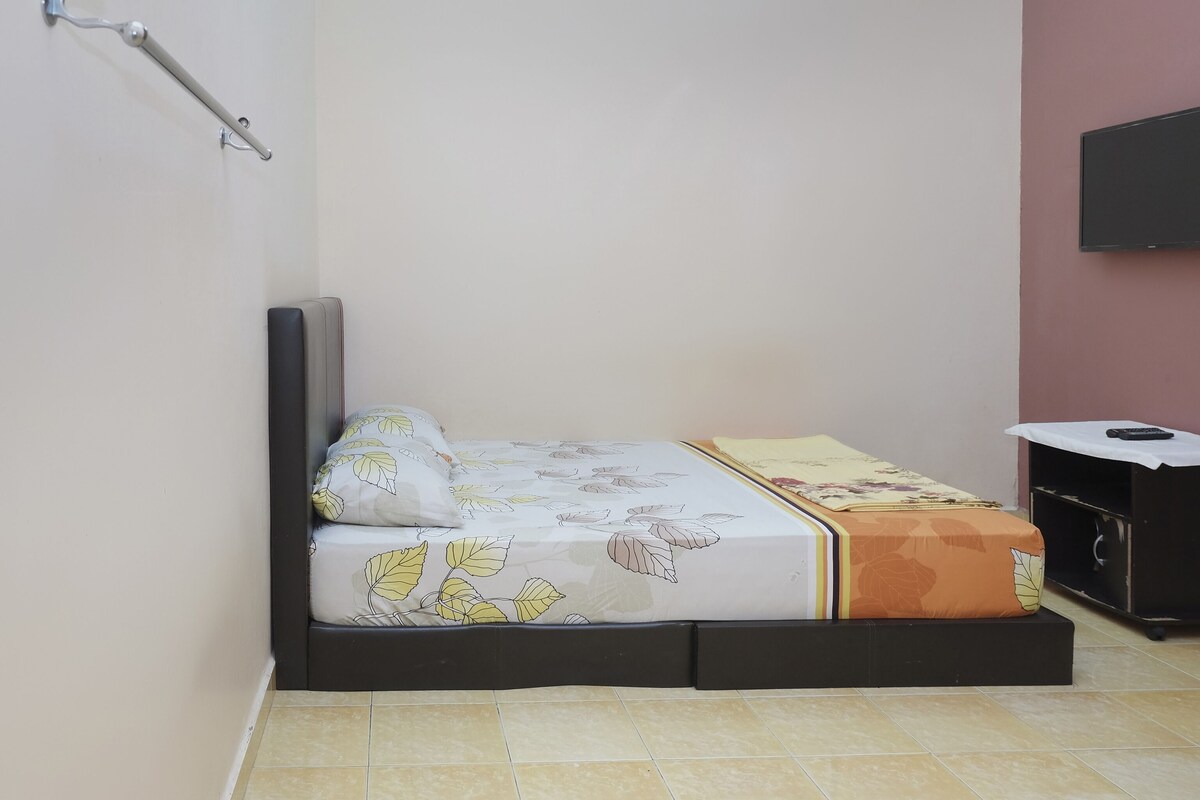 A small, single-bed room with a mattress featuring leaf patterns, a bedside table with a remote control, and a wall-mounted TV.