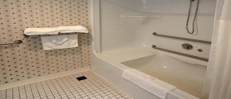 Une salle de bain avec une grande baignoire, une douche et des toilettes. Des serviettes sont accrochées au mur et le sol est recouvert de carreaux disposés en motif quadrillé.