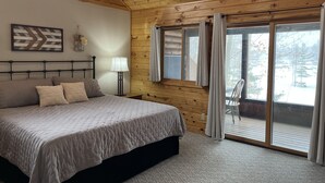 Ein Schlafzimmer mit Holzboden, einem Bett mit Kopfteil, einem Nachttisch, einer Lampe und einem Fenster mit Vorhängen.