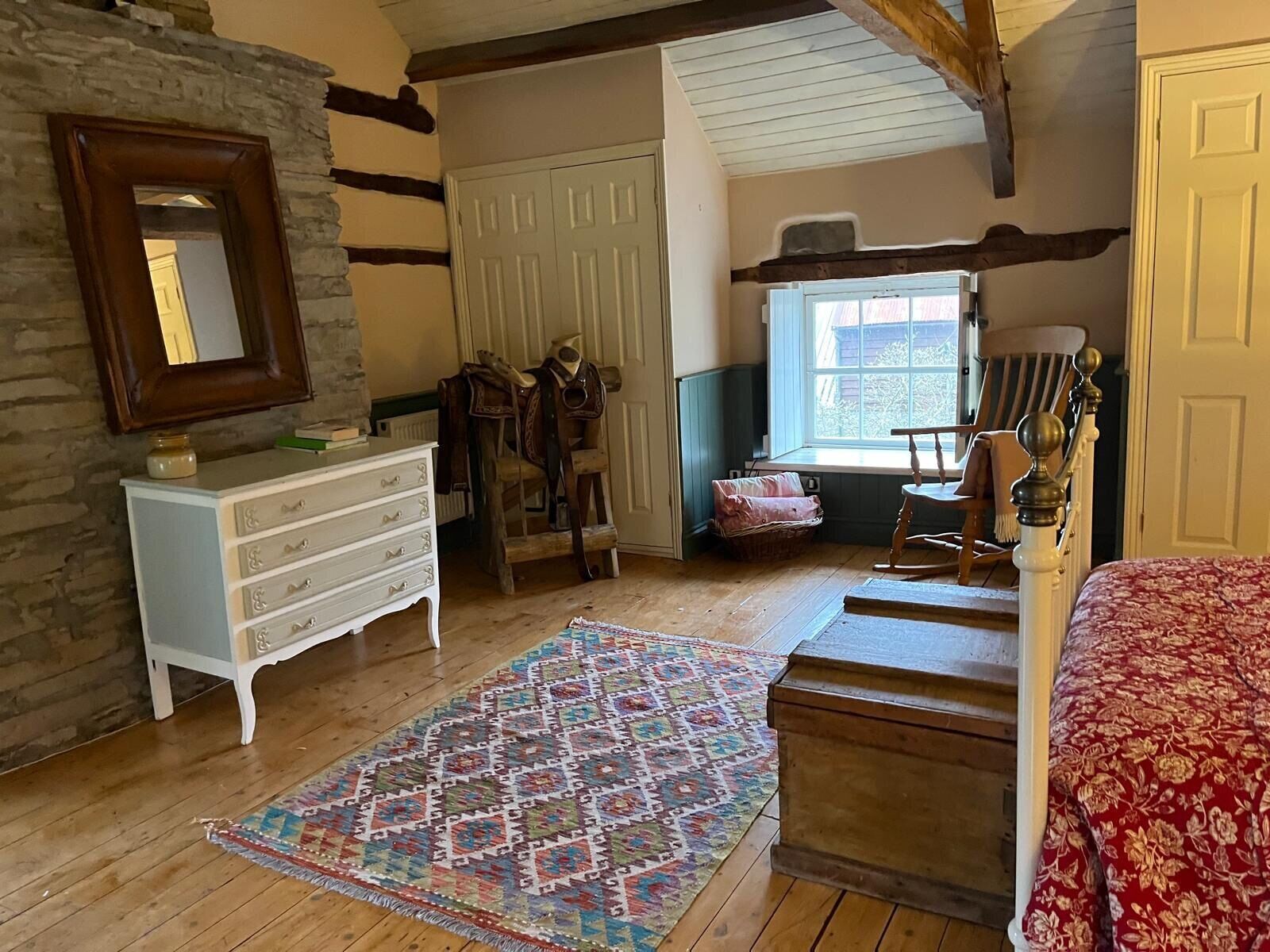 A bedroom with a stone wall, wooden beams, a dresser, a rocking chair, a window with a view, and a patterned rug.