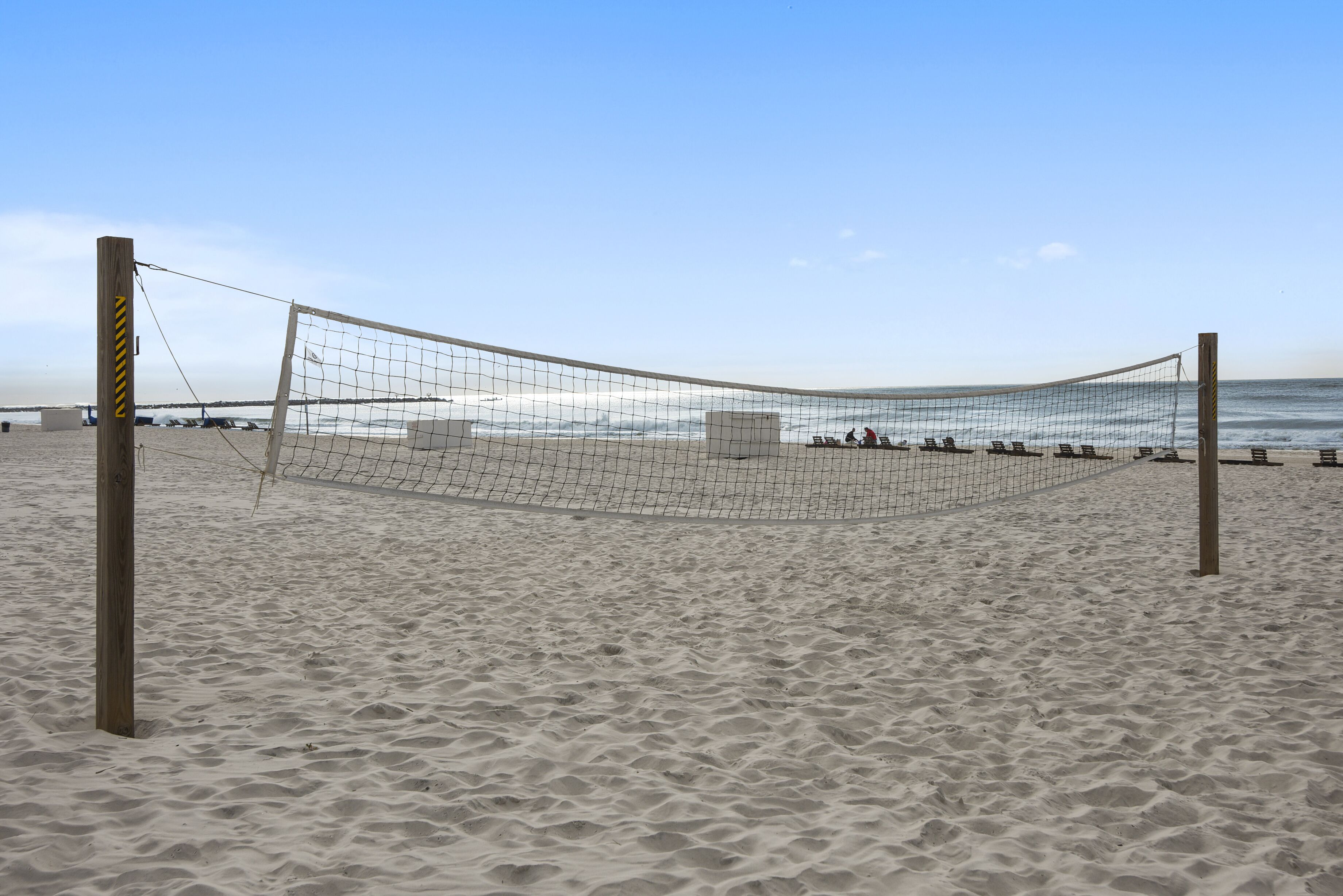 A beach volleyball net with a wooden post and netting, set on a sandy beach with the ocean in the background.