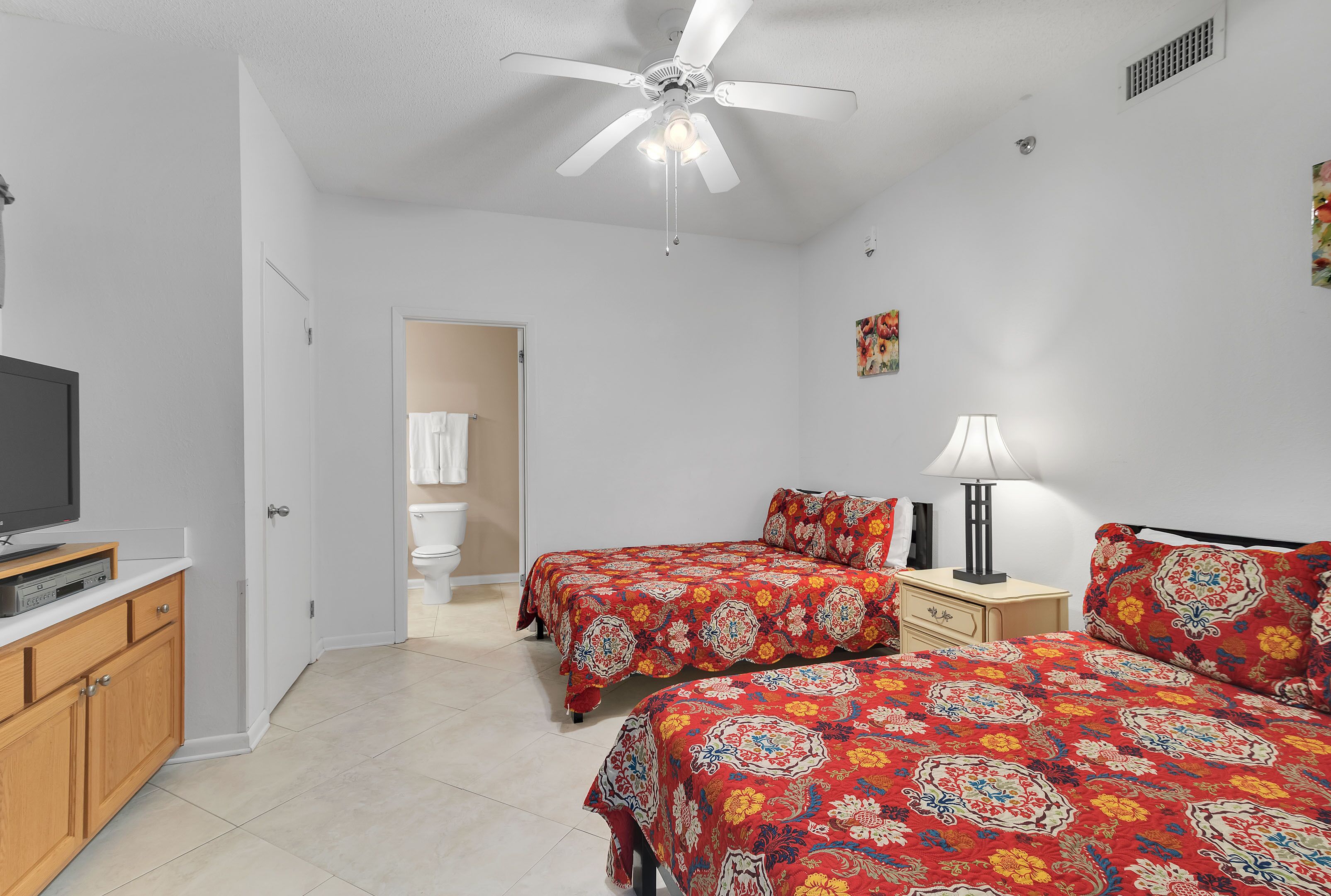 A bedroom with two beds, a ceiling fan, a TV, and a bedside table with a lamp.