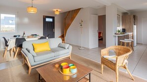 Een moderne woonkamer met een bank, salontafel en een rotan fauteuil. Een trap leidt naar een hoger niveau, een eetgedeelte met tafel en stoelen, en een keuken met een aanrecht en krukken.