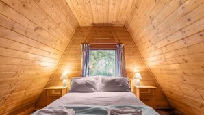 A cozy attic bedroom with wooden walls, a bed with pillows, two bedside lamps, and a window with curtains.