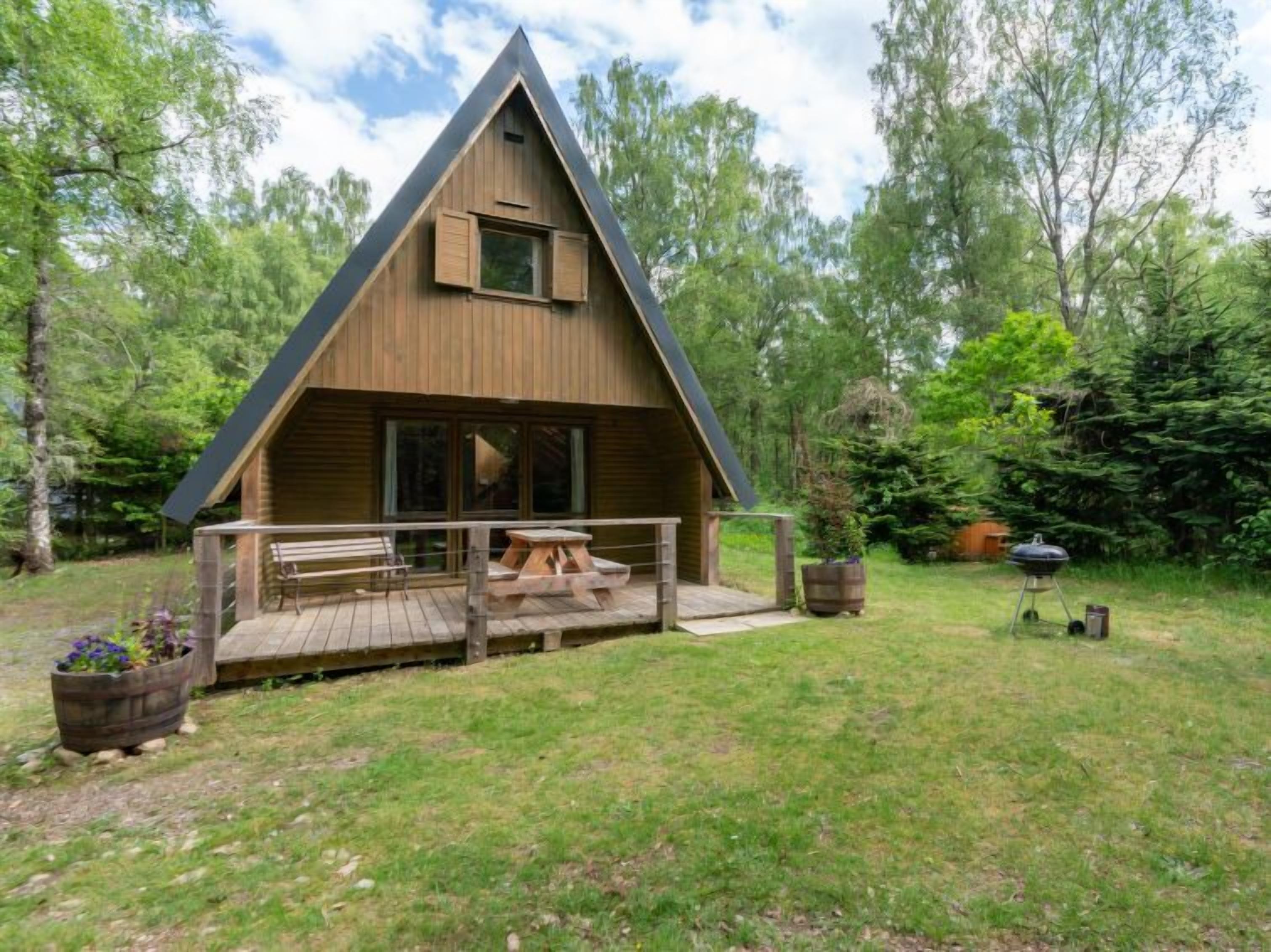 A wooden cabin with a triangular roof, a porch with benches, and a grill in the yard.
