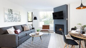 A modern living room with a grey sofa, a round coffee table, a fireplace, and a dining area with a table set for two.