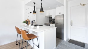A modern kitchen with a central island, bar stools, pendant lights, a clock, and a stainless steel refrigerator.