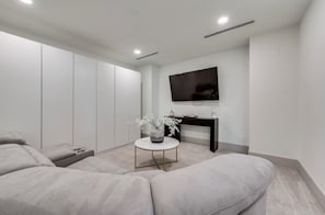 Een moderne woonkamer met een hoekbank, een ronde salontafel met een vaas, een flatscreen televisie aan de wand en een donkere console tafel.