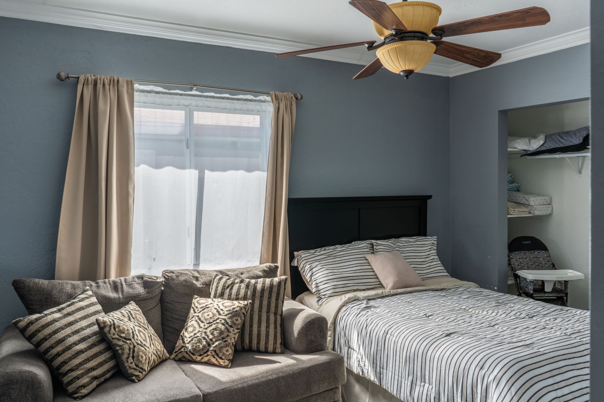 Un dormitorio con una cama, un sofá, un ventilador de techo y una ventana con cortinas.