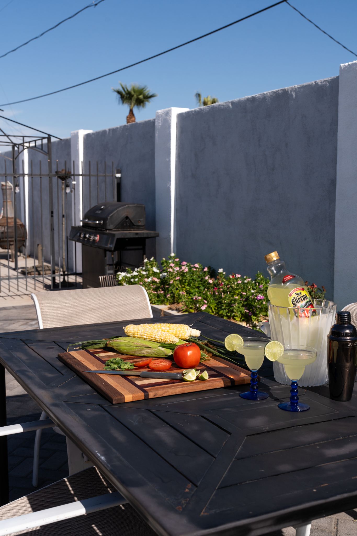 A barbecue setup with a grill, a cutting board with vegetables, and two glasses of a yellow beverage.