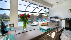 A dining area with a table set for two, a grill, and a view of a swimming pool. - Elegant Modern Home on Salt Water Canal w/ Heated Pool (Cape Coral)