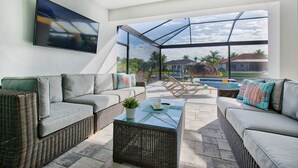 A modern outdoor living space with wicker furniture, a glass-enclosed patio, and a view of a swimming pool and houses. - Elegant Modern Home on Salt Water Canal w/ Heated Pool (Cape Coral)