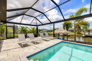 A covered outdoor swimming pool with lounge chairs and a view of a lake and palm trees.