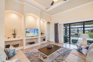 A spacious living room with a large flat-screen TV, comfortable seating, and a view of a pool through a glass door.