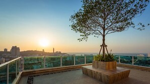 A rooftop terrace with a tree planter, a glass railing, and a cityscape view during sunset.