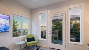 A modern room with a large window, a small round table, a green armchair, and a flat-screen TV displaying a scenic view. - 1stResorts.com Water Views! 2 FAMILY Villas Amalfi/Bellagio on 1 property! (San Diego)
