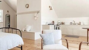 A bedroom with a bed, a chair, and a small table. There is a kitchen area with cabinets and a sink.
